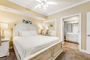 Une chambre à coucher avec un grand lit, un ventilateur de plafond, une commode et une salle de bain visible à travers une porte ouverte.