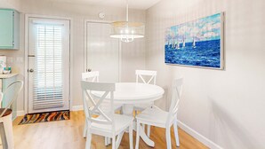 Un'area pranzo con un tavolo rotondo bianco e sedie, una porta bianca con persiane, un lampadario e un quadro con dei velieri al muro.