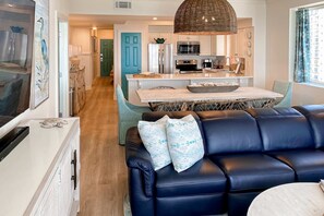 A modern living room with a dark blue leather sofa, a dining area with a wooden table, and a kitchen with white cabinets and a blue door. - Majestic Sun A1101 | Beachfront Condo with Views! (Miramar Beach)