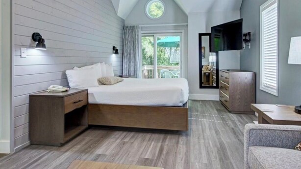 A modern bedroom with a loft bed, a TV, a desk, and a balcony with a view of greenery.