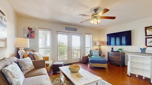 A spacious living room with a sofa, coffee table, and a ceiling fan. A sign on the wall reads "BEACH HOUSE." - Fourth-floor, Caribbean corner condo with ocean views, pool, & hot tub (Santa Rosa Beach)