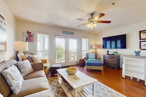 A spacious living room with a sofa, coffee table, and a ceiling fan. A sign on the wall reads "BEACH HOUSE." - Fourth-floor, Caribbean corner condo with ocean views, pool, & hot tub (Santa Rosa Beach)