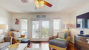A spacious living room with a ceiling fan, a sofa, a chair, a coffee table, and a television. - Fourth-floor, Caribbean corner condo with ocean views, pool, & hot tub (Santa Rosa Beach)