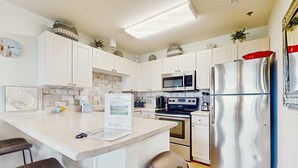 A modern kitchen with white cabinets, stainless steel appliances, a central island with bar stools, and a trash bin. - Fourth-floor, Caribbean corner condo with ocean views, pool, & hot tub (Santa Rosa Beach)