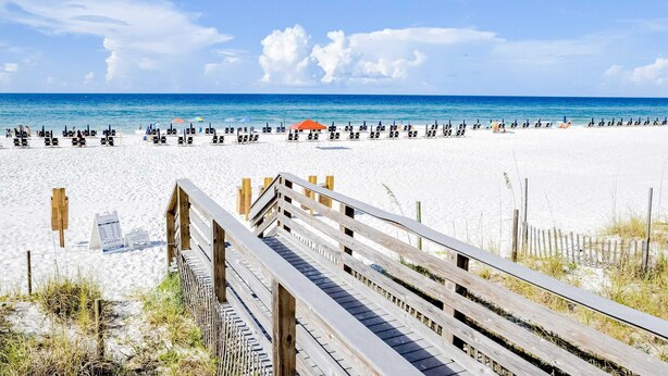 Ein Strand mit einer Holzpromenade, die zum Wasser führt, weißem Sand und Strandstühlen.