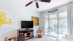 A living room with a flat-screen TV, wooden entertainment center, wicker chair, and a sofa. - Beautiful gem at Bayou Village, golf cart, WiFi, & resort pools (Miramar Beach)