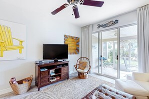 A living room with a flat-screen TV, wooden entertainment center, wicker chair, and a sofa. - Beautiful gem at Bayou Village, golf cart, WiFi, & resort pools (Miramar Beach)