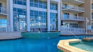 Uma piscina com deck ladrilhado, uma fonte de água e um edifício ao fundo com janelas amplas e varandas.