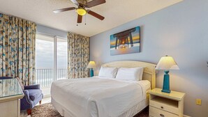 Quarto com cama, duas mesas de cabeceira com luminárias, ventilador de teto, janela com cortinas e um quadro na parede.