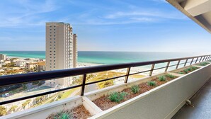 A balcony with a view of the beach and ocean, a tall building, and a road with cars. - Gulf-view resort condo with pools, hot tubs, balcony, WiFi, W&D, & AC (Miramar Beach)