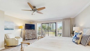 Ein Hotelzimmer mit einem großen Bett, einem Deckenventilator, einem Fernseher, einem Schminktisch, einem Sessel und einem Balkon mit Blick auf Palmen.