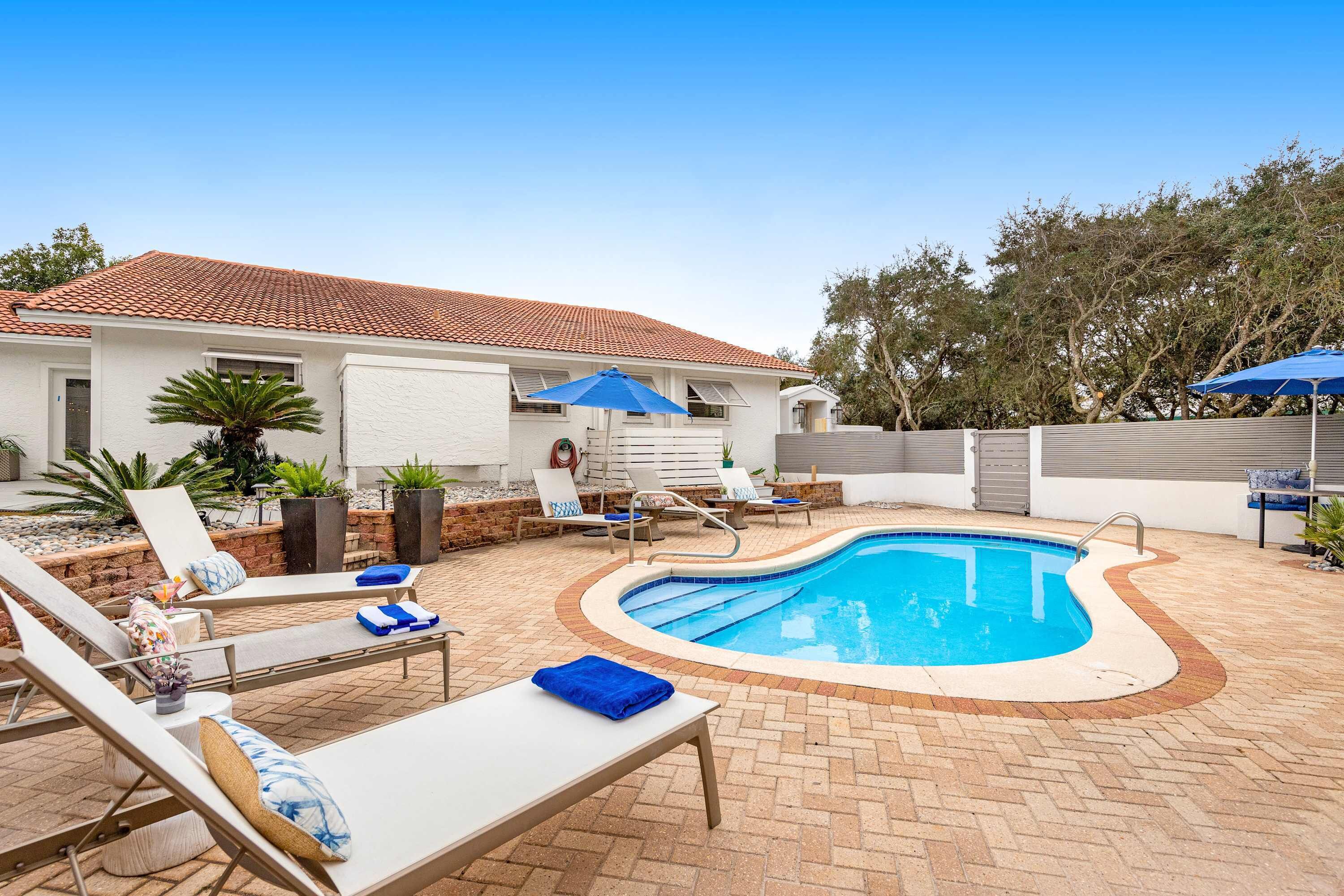 A swimming pool with lounge chairs and umbrellas, surrounded by a brick patio and a white building with a tiled roof.