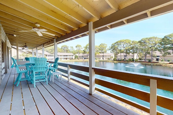 A covered outdoor area with wooden flooring, turquoise chairs, and a view of a body of water with houses in the background. - Lakefront condo with on-site golf, pool access, tennis, gym, & balcony (Miramar Beach)