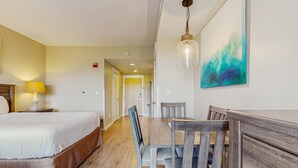 Une chambre d’hôtel avec un lit, une table à manger en bois avec des chaises, un tableau au mur et un couloir menant à une autre pièce.