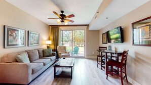 A spacious living room with a sofa, coffee table, and dining table. There is a ceiling fan, a TV mounted on the wall, and a large window providing natural light. - LaSata beauty with patio, pools, hot tubs, fitness center, AC & beach access (Miramar Beach)