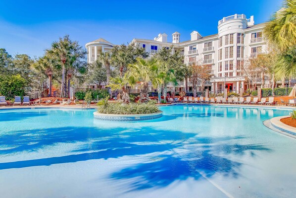 A large pool surrounded by palm trees and a hotel with multiple balconies and a tower.