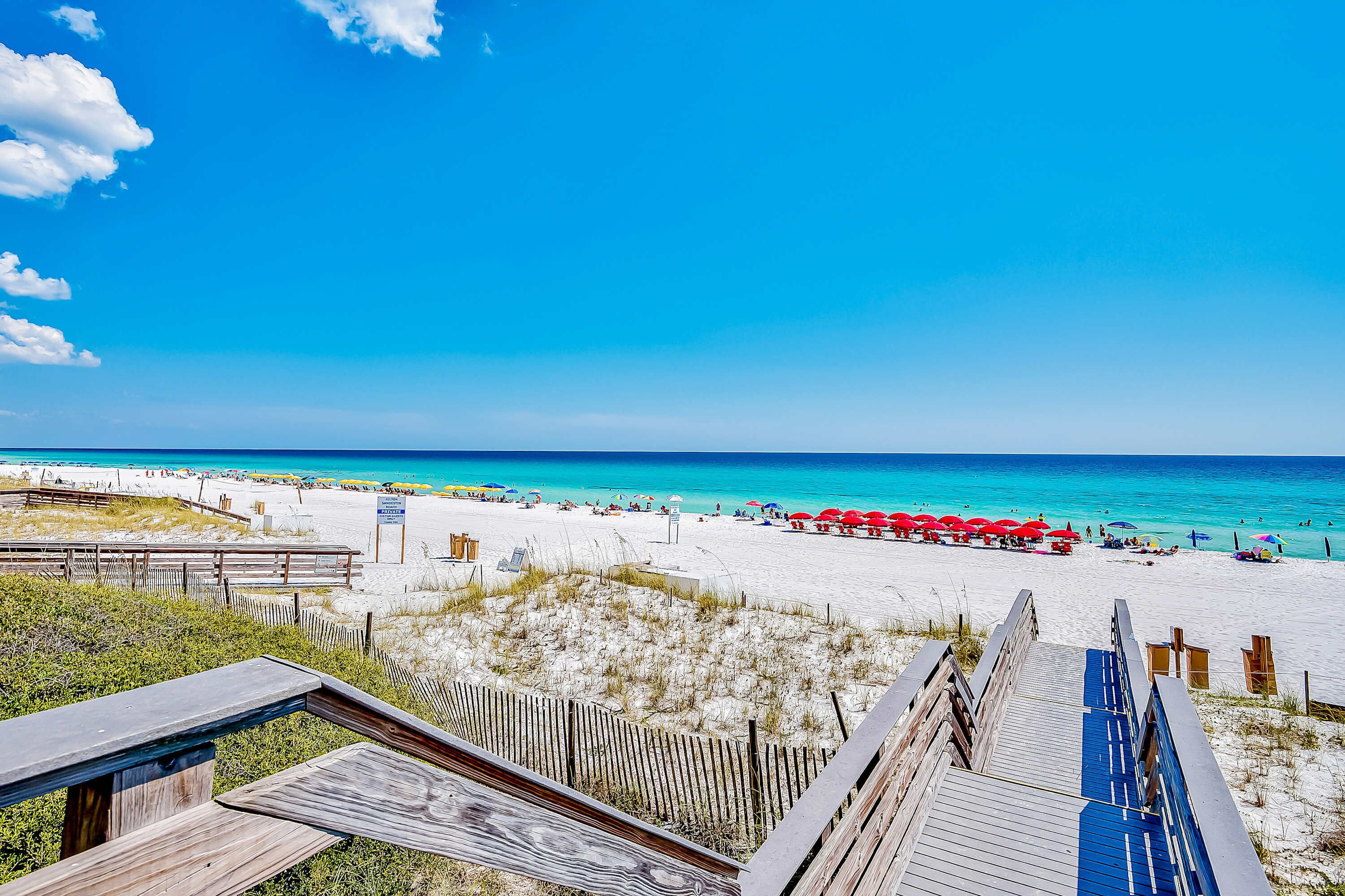 Une plage avec une promenade en bois menant au rivage, une plage de sable et une eau d’un bleu limpide.