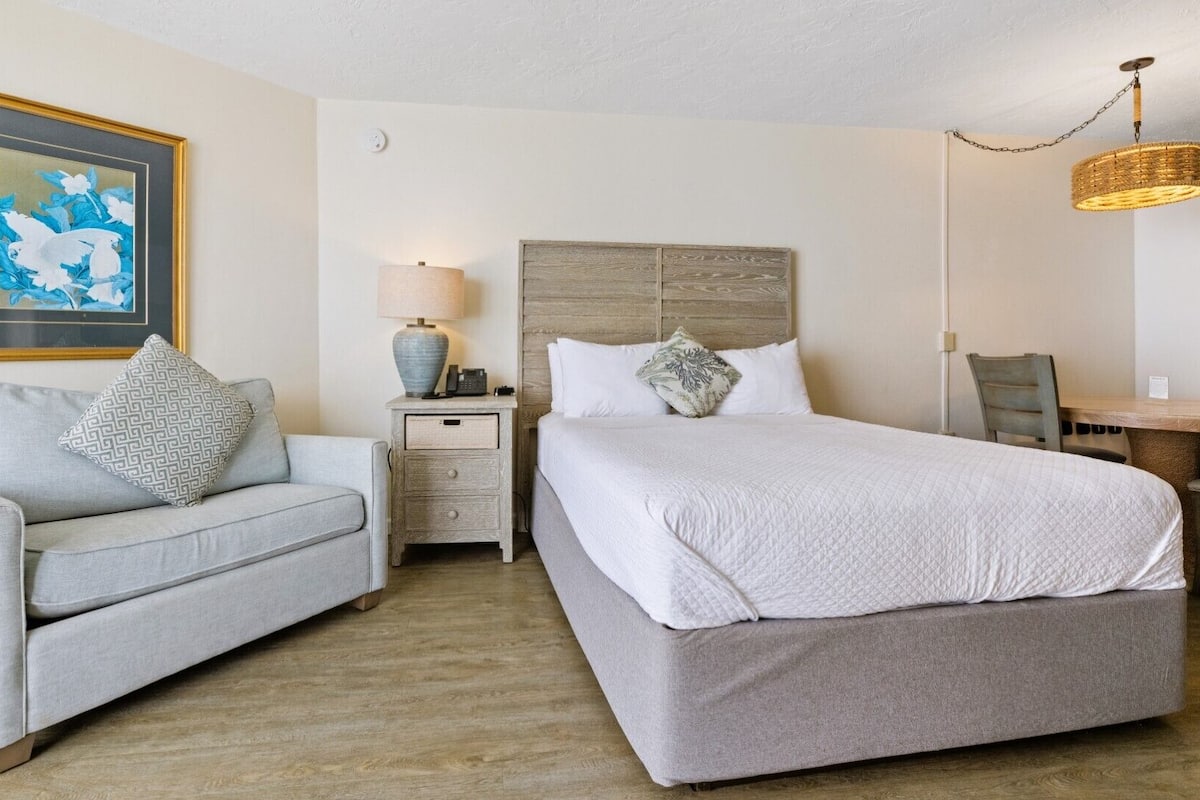 A hotel room with a bed, a sofa, a nightstand, a lamp, a framed picture, and a wicker hanging light fixture.