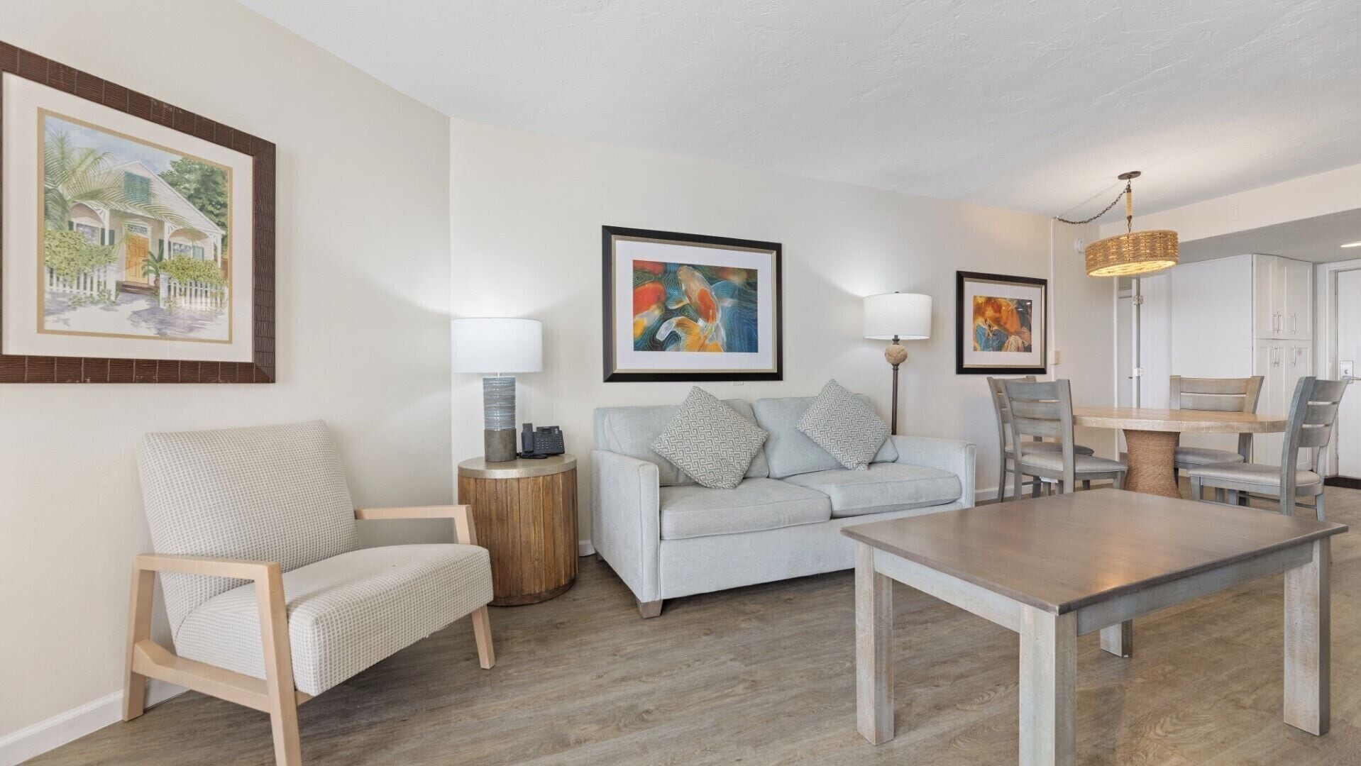 A living room with a sofa, armchair, and dining table. There are framed artworks on the walls and a chandelier.