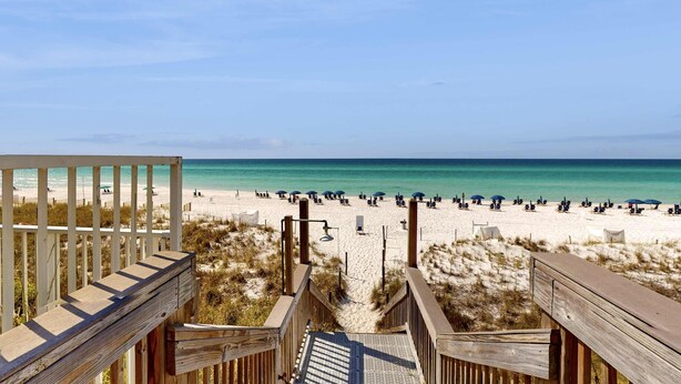 Ein Holzsteg führt zu einem Strand mit Menschen und Sonnenschirmen.