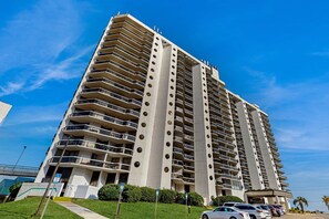 A high-rise building with multiple balconies, a parking area, and a clear blue sky. - 11th-floor home w/free WiFi, washer/dryer, ocean views, & shared hot tub & pool (Miramar Beach)