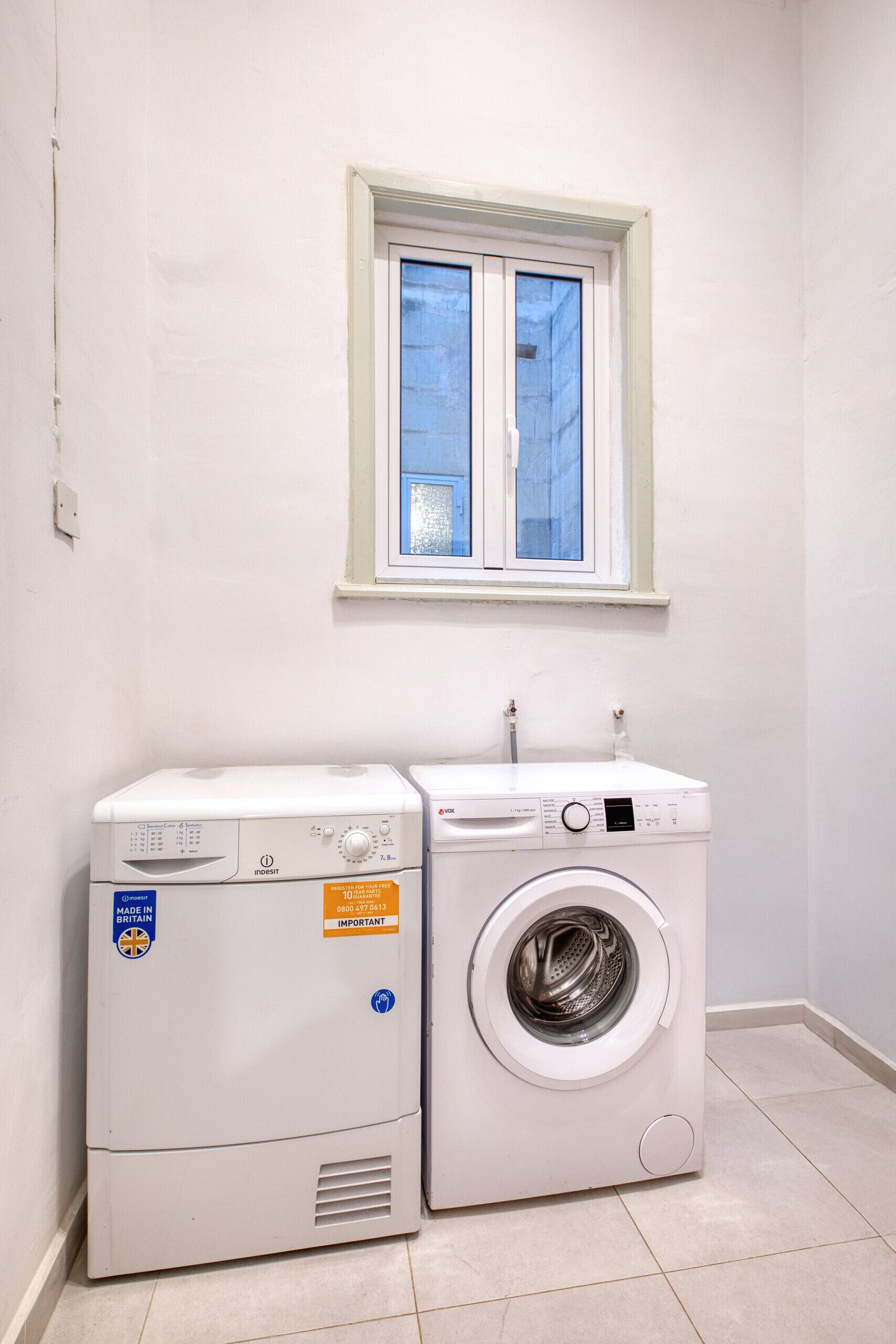 Un área de lavandería con lavadora de carga frontal y secadora de carga superior, una ventana sobre los electrodomésticos y piso de baldosas.