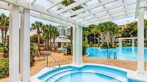 A pool area with a pergola, palm trees, and a building in the background. - First-floor condo with a kitchen, patio, pool, hot tub, grill, & spa access (Miramar Beach)