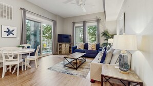A spacious living room with a sofa, coffee table, and TV. There is a dining area with a table and chairs. The room has large windows and a ceiling fan. - First-floor condo with a kitchen, patio, pool, hot tub, grill, & spa access (Miramar Beach)