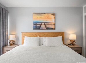 Et hotelværelse med en stor seng, et træhovedgærde, to sengeborde med lamper og et billede af en solnedgang over en moler.