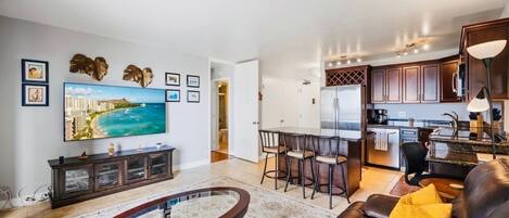 A modern living room with a TV, a coffee table, and a sofa. There is a kitchen area with cabinets and a dining table.