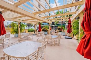 En udendørs patio med runde borde og fletstole, overdækket af en træpergola med røde parasoller.