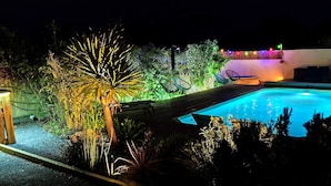 Ein Pool, umgeben von Pflanzen und Stühlen, mit einer Holzterrasse und einem Zaun.