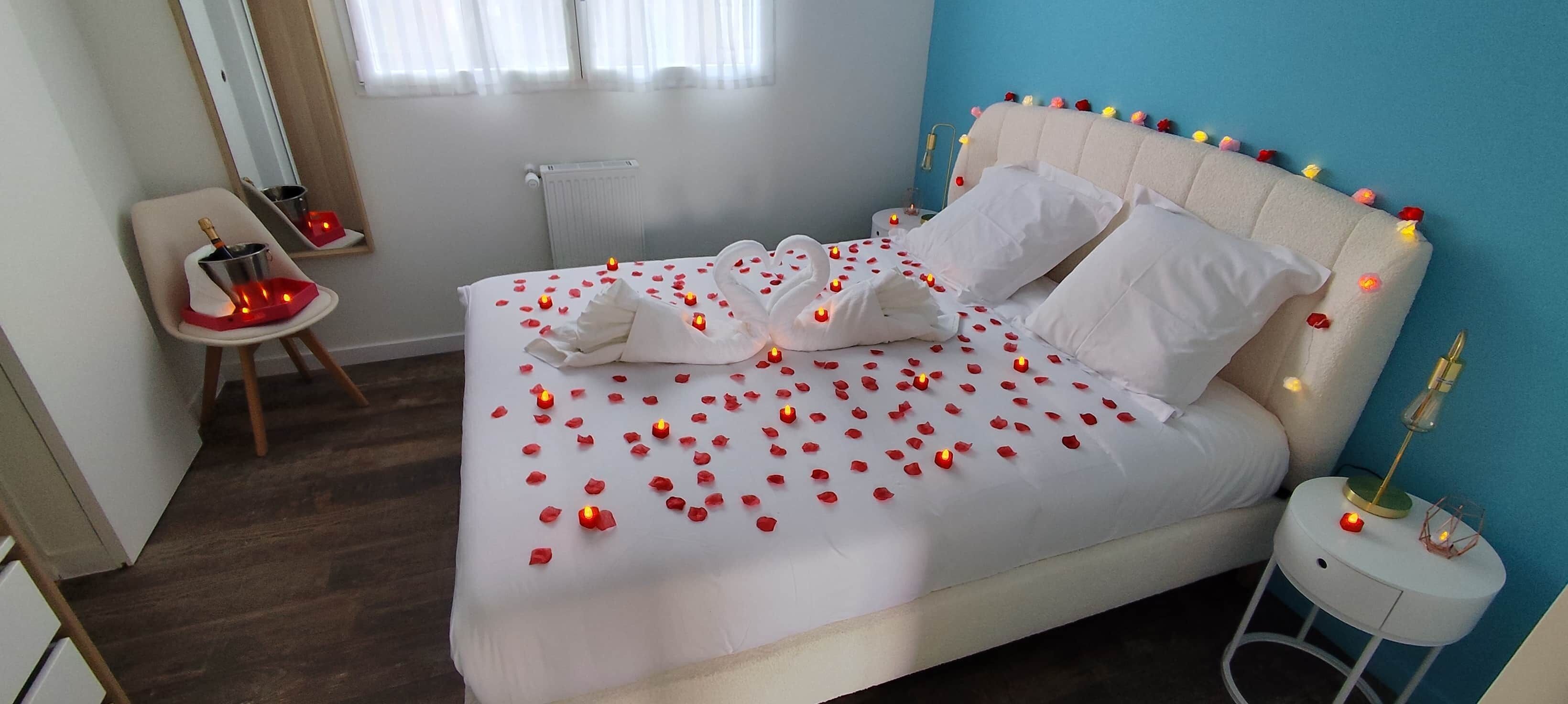 A bed with rose petals and candles, a chair with a bottle, and a bedside table with a lamp.
