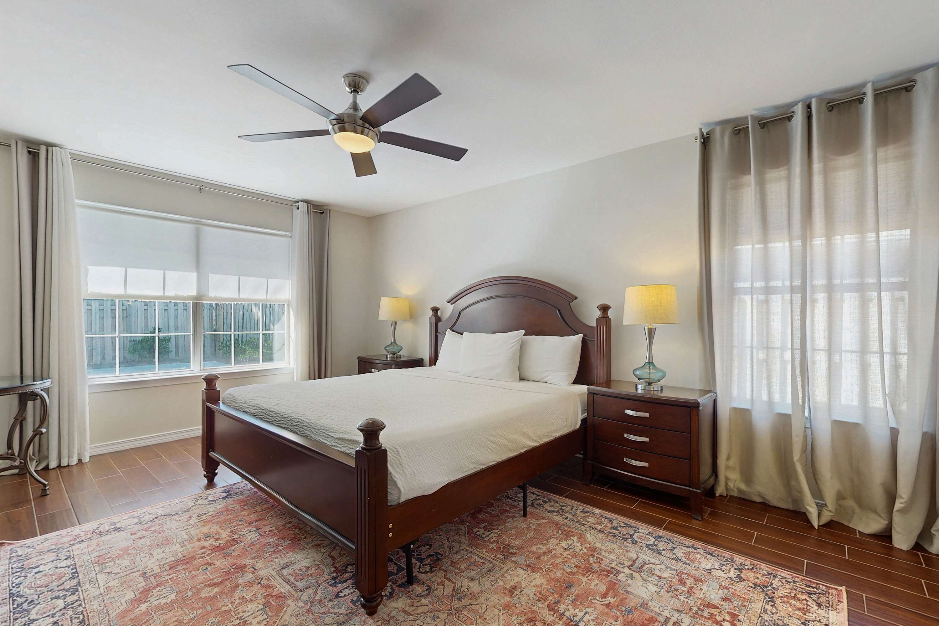 A bedroom with a wooden bed, two bedside tables with lamps, a ceiling fan, a large window with curtains, and a patterned rug.
