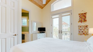 A bedroom with a large bed, a television on a dresser, and a balcony with a view of buildings. - Beachfront dog-friendly home with spacious decks and on-site pool (St. George Island)