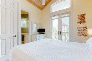 A bedroom with a large bed, a television on a dresser, and a balcony with a view of buildings. - Beachfront dog-friendly home with spacious decks and on-site pool (St. George Island)