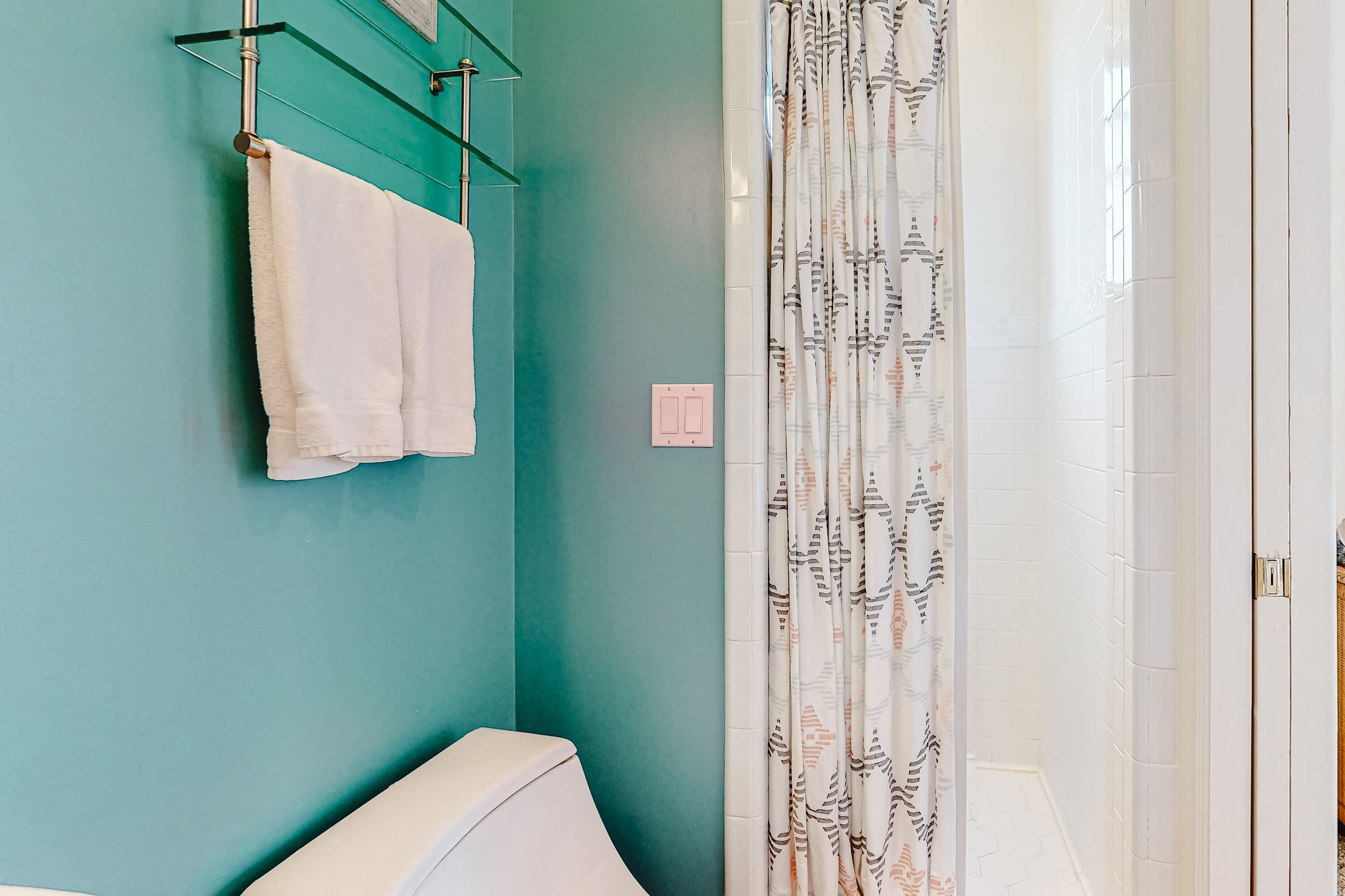 A bathroom with teal walls, a toilet, a towel rack with two towels, and a shower curtain with a leaf pattern.