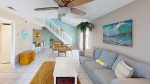 A living room with a ceiling fan, a sofa, a coffee table, a staircase, and a painting of a wave. - Quaint Miramar Beach home with loft, pool, patio, full kitchen, & washer/dryer (Miramar Beach)