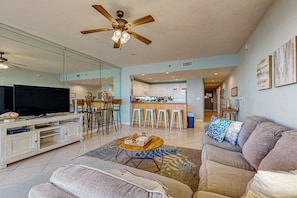 A spacious living room with a large flat-screen TV, a ceiling fan, and a kitchen area in the background. - Beautiful, waterfront condo w a shared pool, gym, & beach access (Destin)