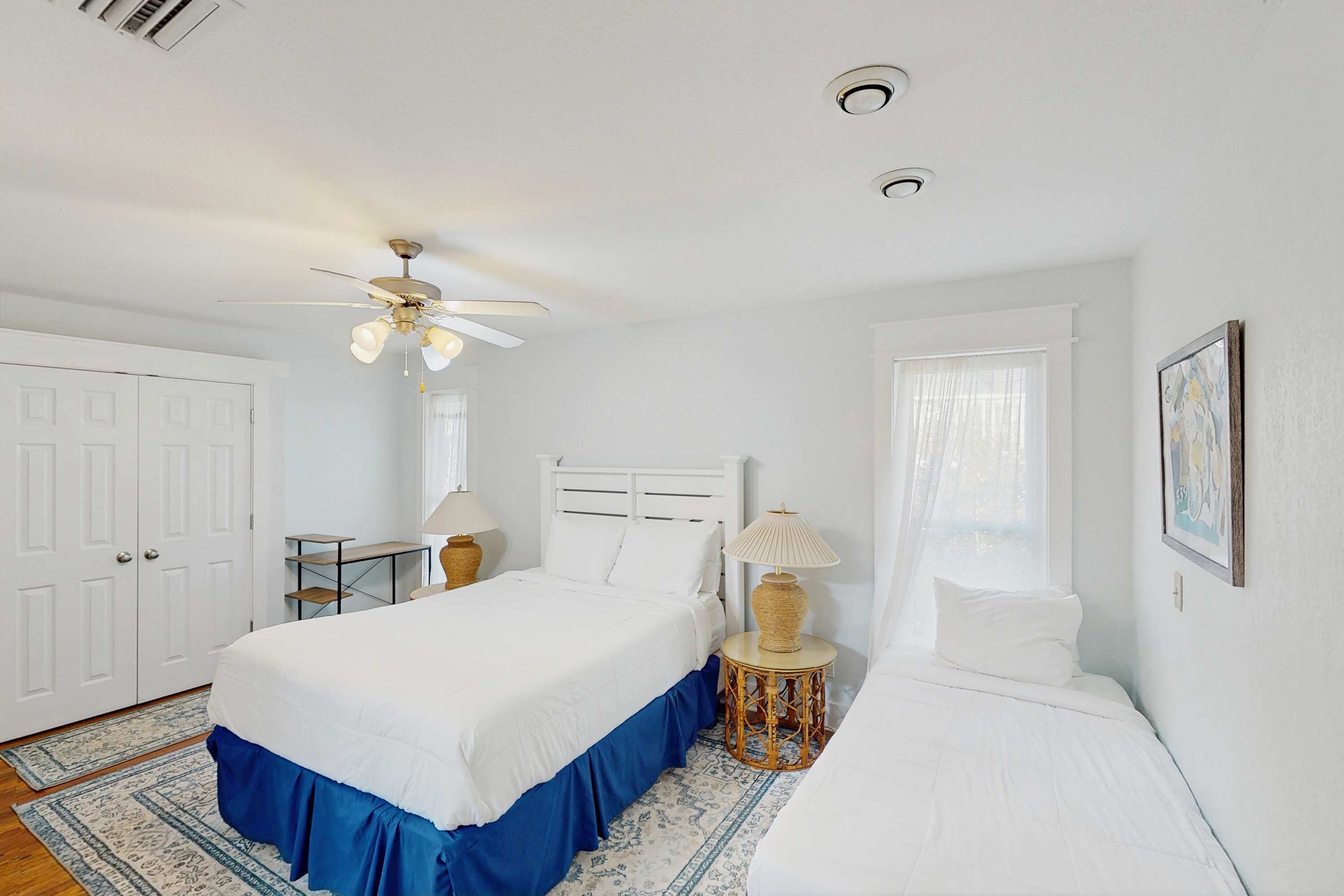 Une chambre avec un grand lit, un lit plus petit, un bureau, une chaise, un ventilateur de plafond et une fenêtre avec des rideaux.