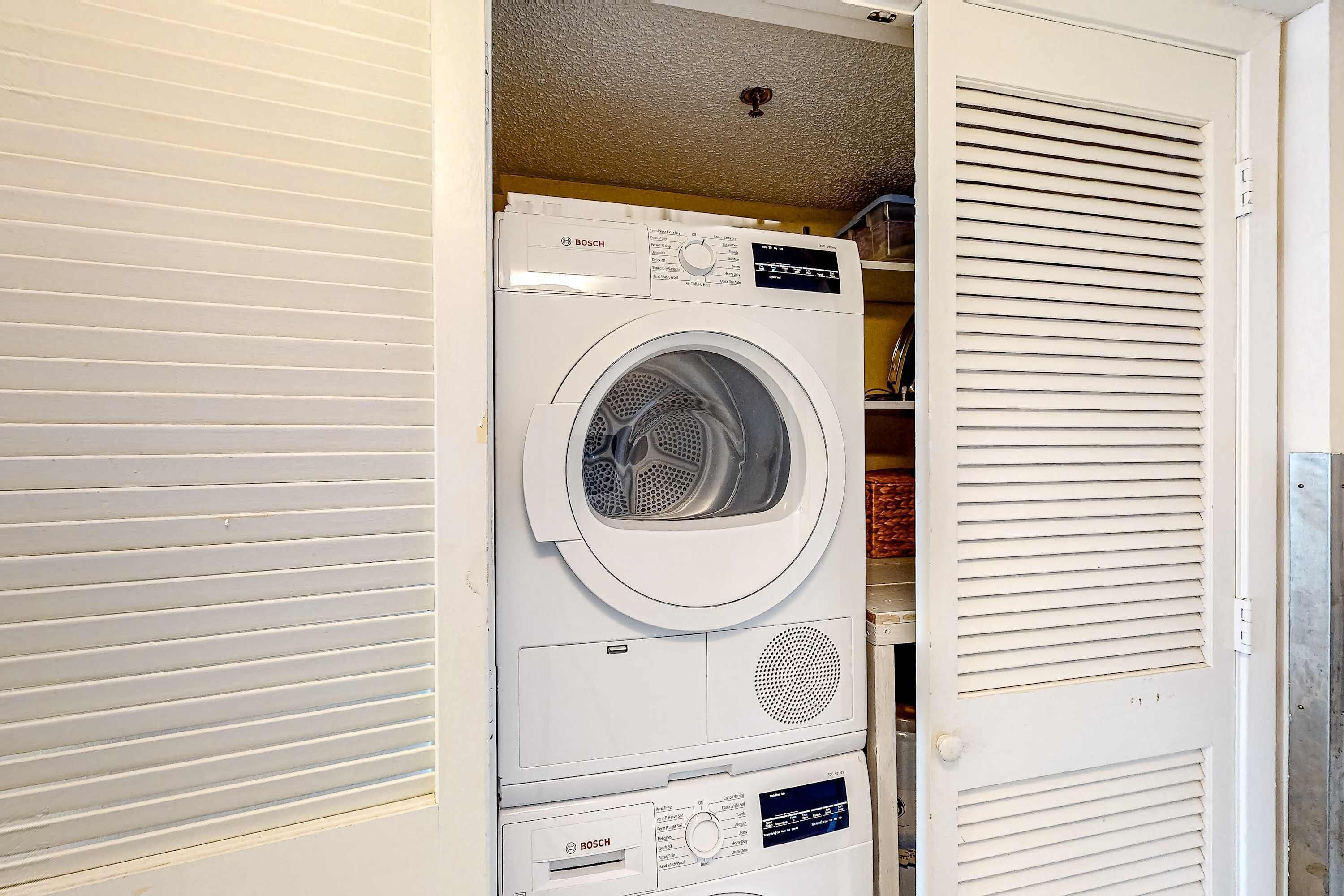 Une machine à laver Bosch blanche et compacte, installée dans un petit local à buanderie avec une porte fermée et une bouche d’aération au plafond.