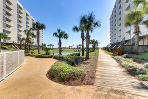 A pathway flanked by palm trees and a water body, leading to a multi-story building with balconies. - Gulf-view condo with balcony, pools, hot tub, beach access (Miramar Beach)