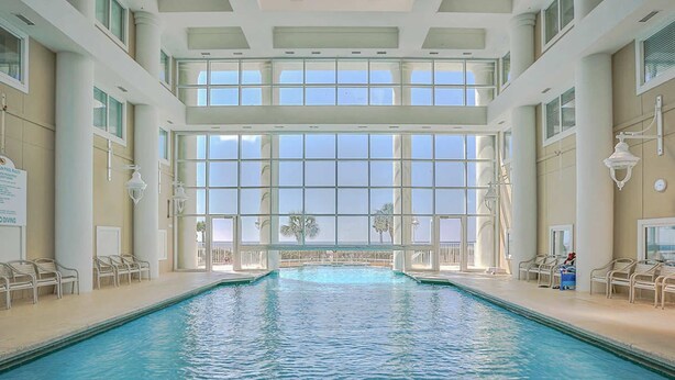 An indoor swimming pool with clear glass windows, lounge chairs, and a view of palm trees. - Majestic Sun A912 | Beachfront Condo with Views! (Miramar Beach)