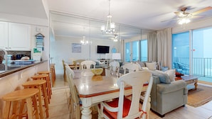 A spacious living area with a dining table, chairs, and a sofa. There is a kitchen area with a counter and stools. A ceiling fan and a TV are visible. - Majestic Sun A912 | Beachfront Condo with Views! (Miramar Beach)