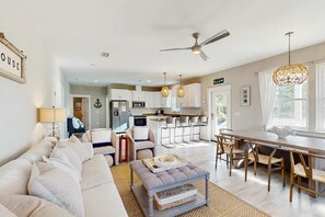 A modern living room with a sofa, ottoman, and dining area with chairs. There is a kitchen in the background with white cabinets and a bar area. - 2 King + bunk room beach retreat with open layout, AC, private heated pool, fire pit & grill, 5 minute walk to beach (Port St. Joe)