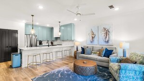 Eine moderne Küche mit hellblauen Schränken, einer weißen Arbeitsplatte und einem Edelstahl-Kühlschrank. Es gibt eine Bar mit Hockern und einem runden Couchtisch. Das Wohnzimmer verfügt über ein graues Sofa mit gemusterten Kissen, einen blau gemusterten Sessel und einen blauen Teppich.