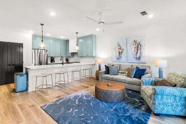 Una cocina moderna con armarios azul claro, una encimera blanca y un refrigerador de acero inoxidable. Hay una zona de bar con taburetes y una mesa de centro redonda. La sala de estar tiene un sofá gris con cojines estampados, un sillón azul con estampado y una alfombra azul.