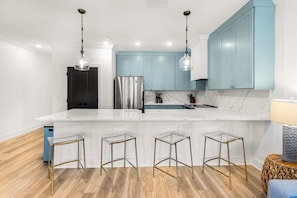 Una cocina moderna con encimera de mármol blanco, electrodomésticos de acero inoxidable y armarios azul claro. Hay tres taburetes transparentes con marcos dorados y una mesita de noche de fibra natural.