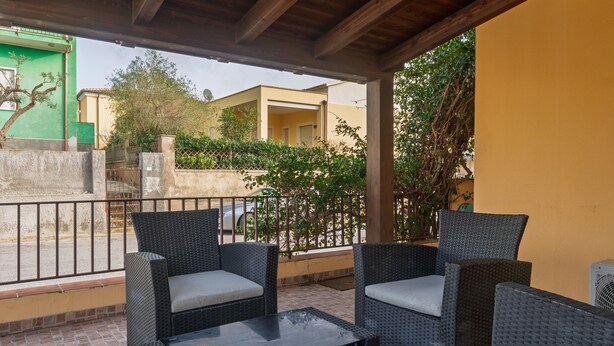 A covered outdoor seating area with wicker furniture and a glass table, overlooking a street with buildings and greenery. - Holiday Home 'La Casa Del Bouganville' Near Beaches with Courtyard & Air Conditioning (Golfo Aranci)