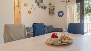 A dining area with a table set for two, a red teapot, a coffee pot, and a decorative jar. There are chairs, a sofa, and a wall with clocks and a shelf with plants. - Holiday Home 'La Casa Del Bouganville' Near Beaches with Courtyard & Air Conditioning (Golfo Aranci)